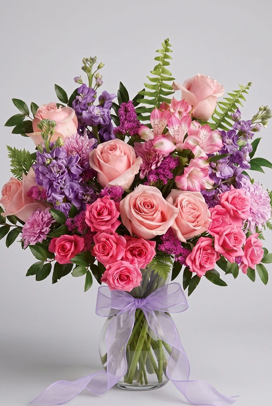 Beautiful Spirit arrangement of pink roses, hot pink spray roses, lavender stock, pink alstroemeria and magenta statice in a clear glass vase with a lavender bow