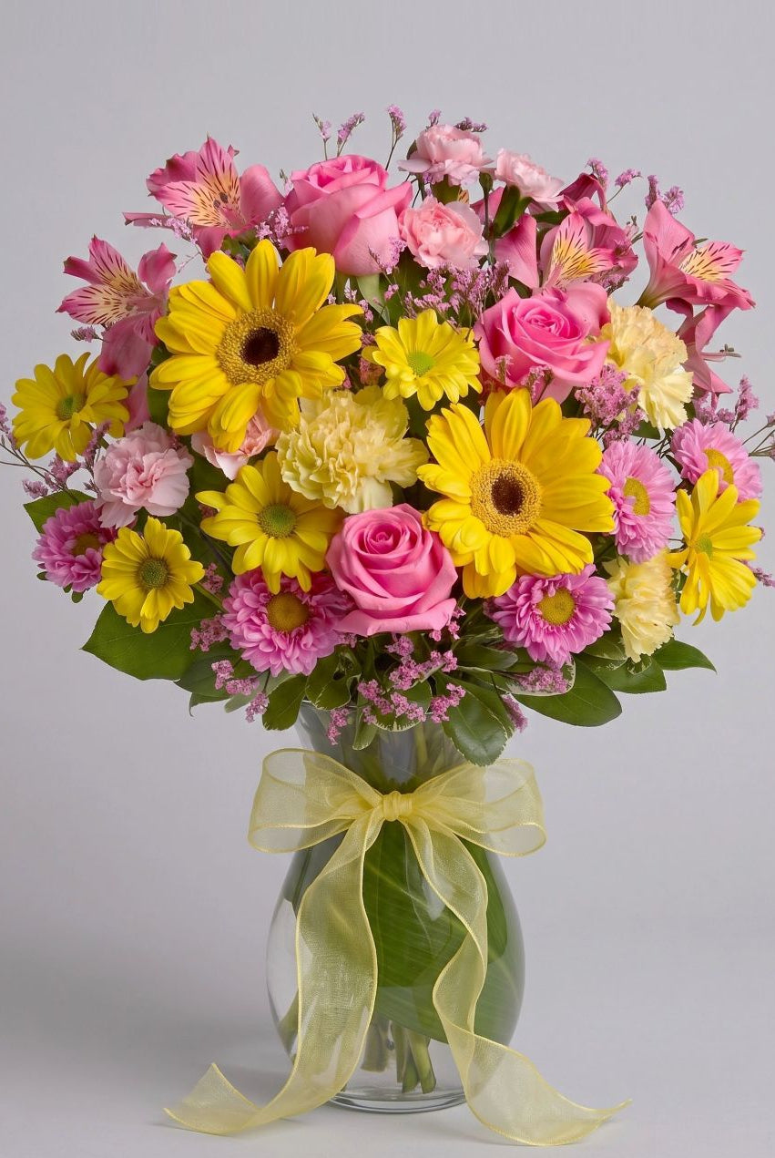 Dazzling Day arrangement of yellow gerbera daisies, pink roses, pink alstroemeria, carnations and asters in a leaf-lined glass vase with a yellow bow