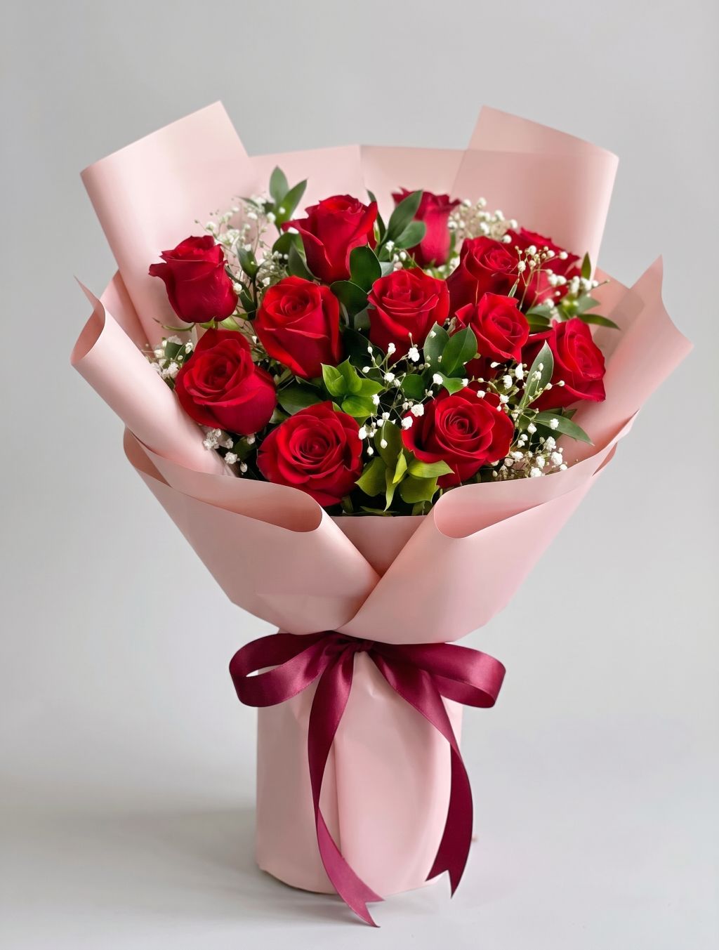 Deluxe Dozen Red Roses Bouquet of twelve red roses with baby’s breath and greenery wrapped in blush pink paper with a burgundy satin ribbon