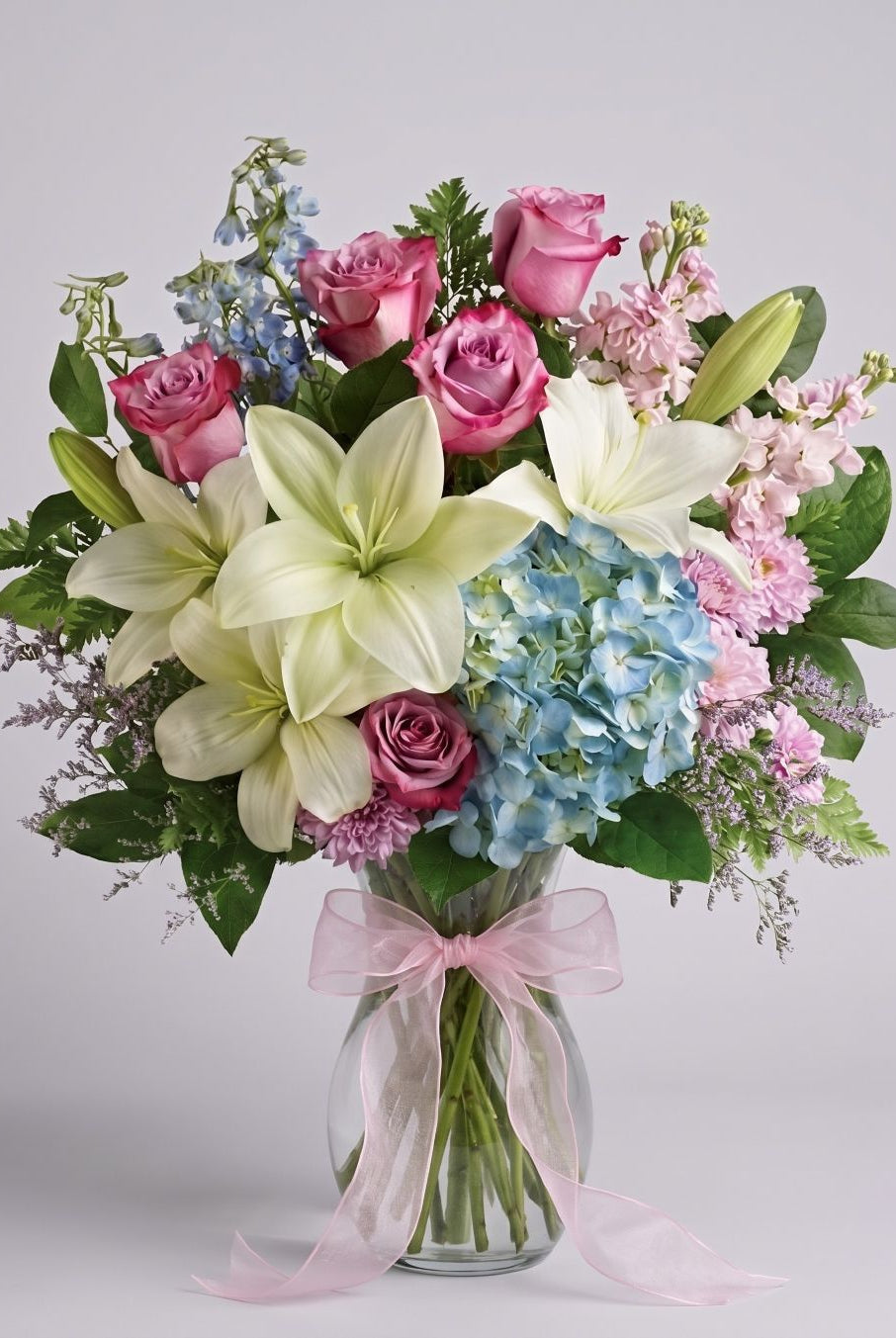 Gentle Comfort arrangement of white lilies, mauve-pink roses, blue hydrangea, blue delphinium, pink stock and purple limonium in a clear glass vase with a pink bow