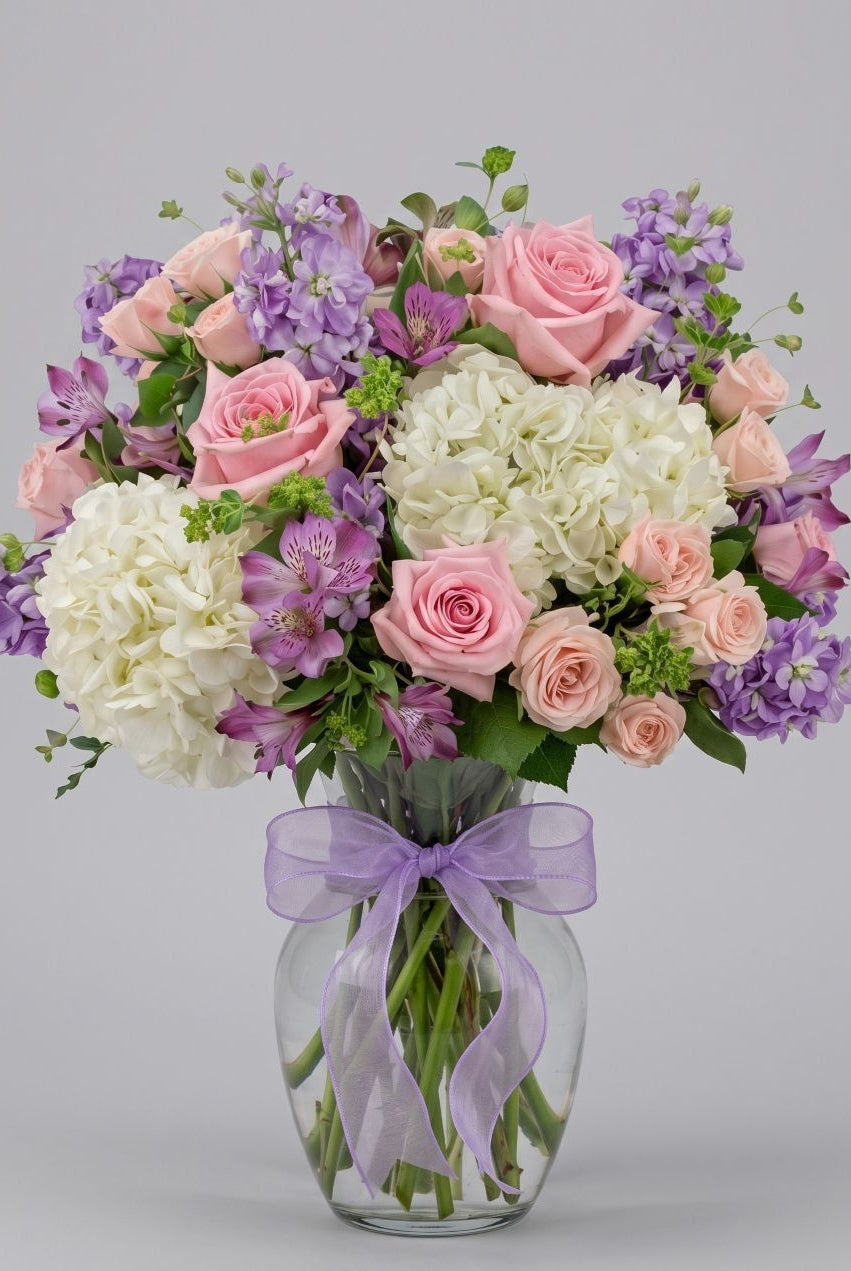 Gentle arrangement of pink roses, peach spray roses, white hydrangea, lavender stock and purple alstroemeria in a clear glass vase with a lavender bow