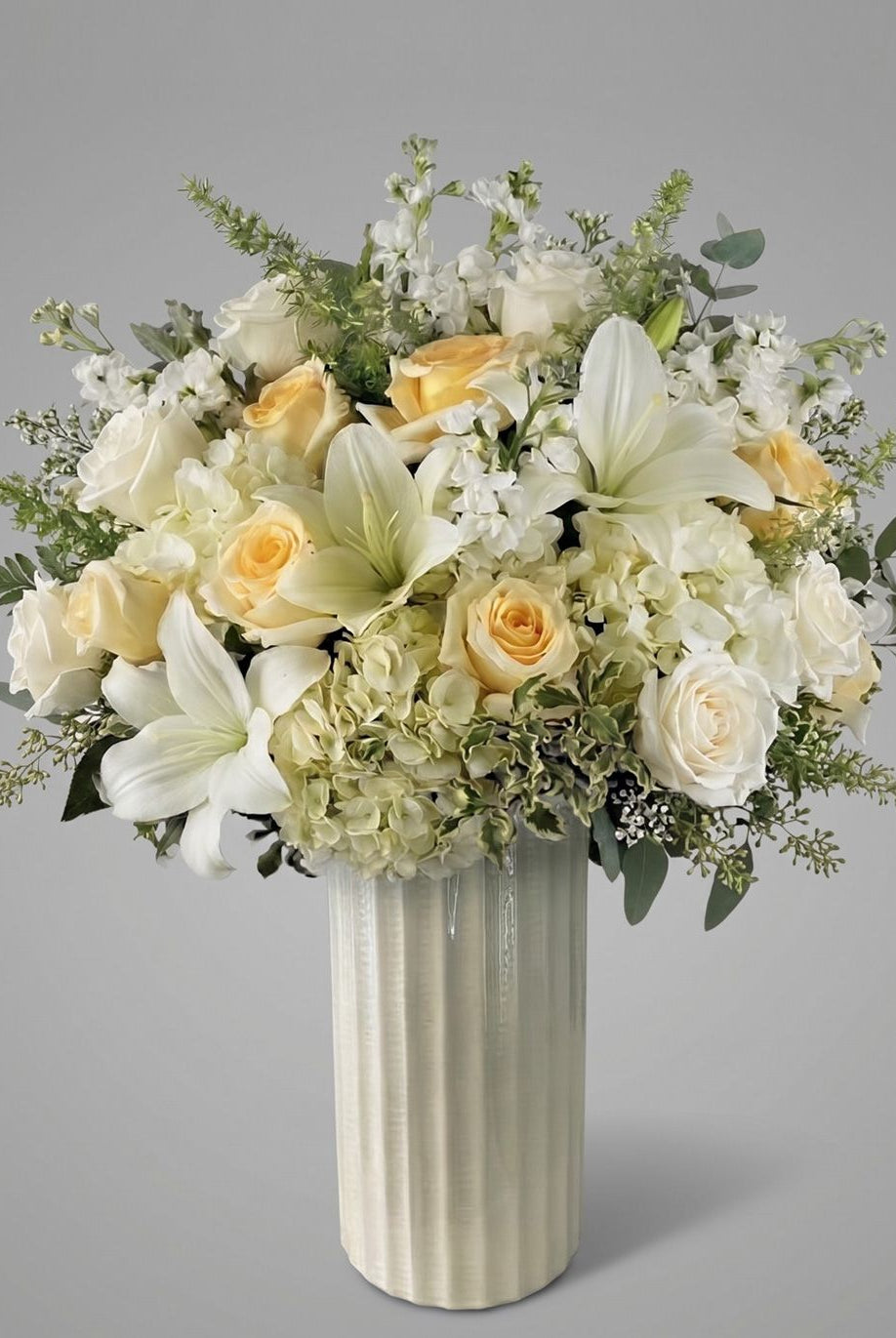 Hope arrangement of cream and peach roses, white lilies, white hydrangea, white stock and eucalyptus in a tall ivory ribbed ceramic vase