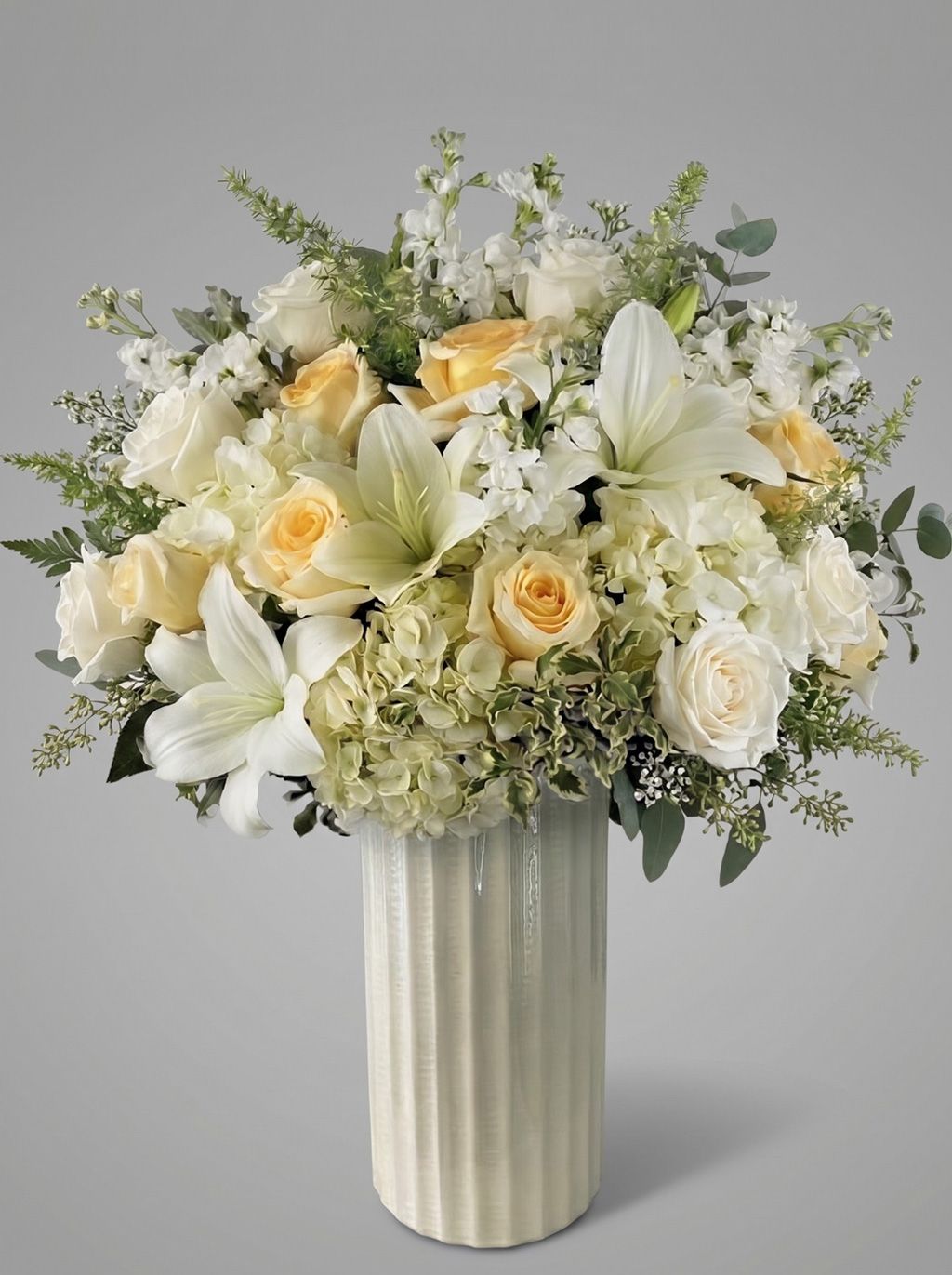 Hope arrangement of cream and peach roses, white lilies, white hydrangea, white stock and eucalyptus in a tall ivory ribbed ceramic vase