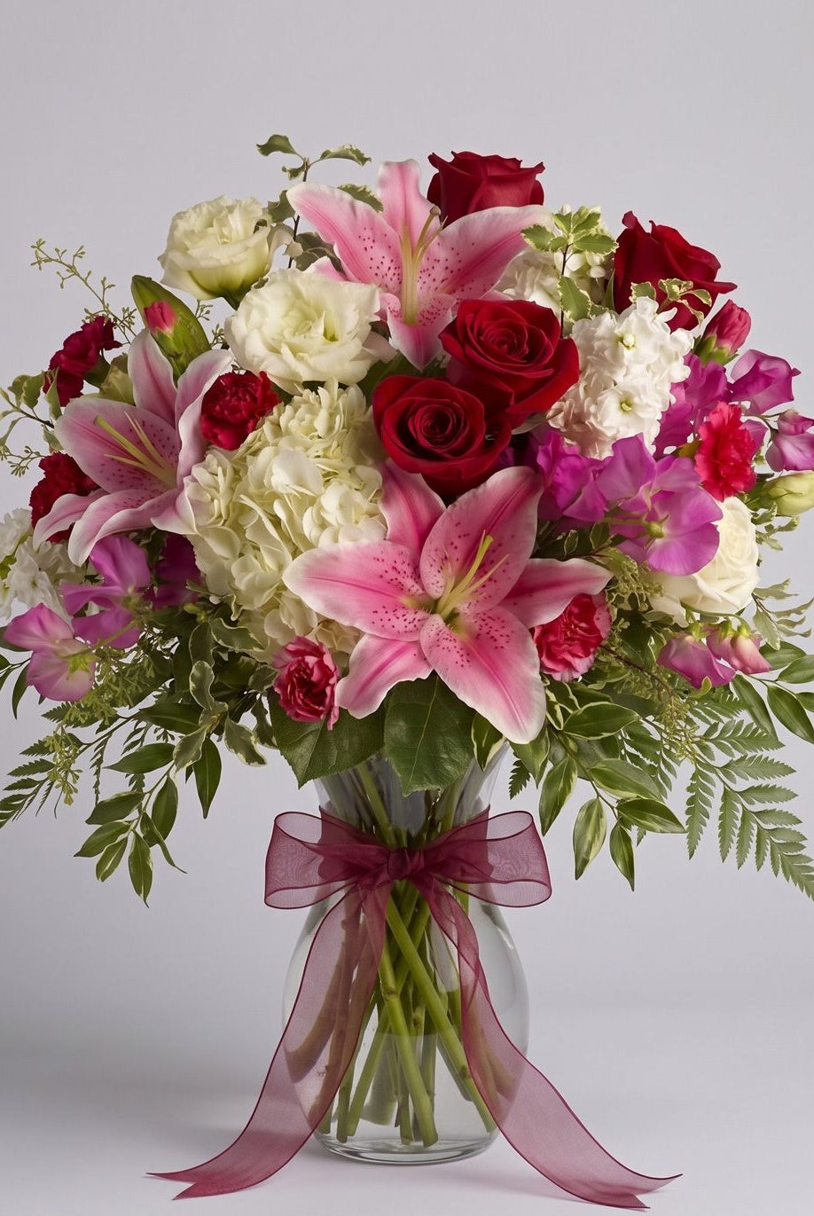 Kensington Gardens arrangement of pink stargazer lilies, red roses, white hydrangea, lisianthus, sweet peas and carnations in a clear glass vase with a burgundy bow.