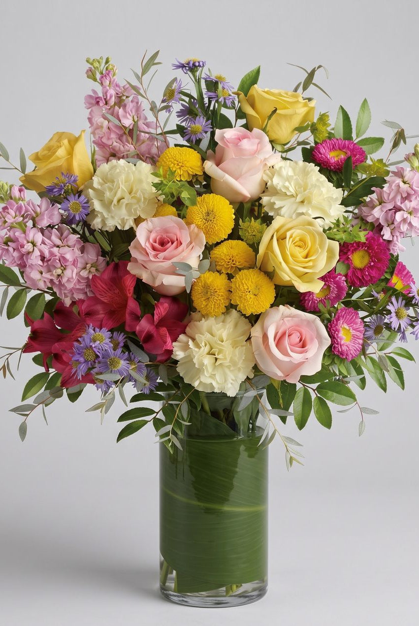 Seasonal spring vase arrangement of pink and yellow roses, cream carnations, yellow button mums, pink stock, red alstroemeria and purple asters in a leaf-lined glass vase