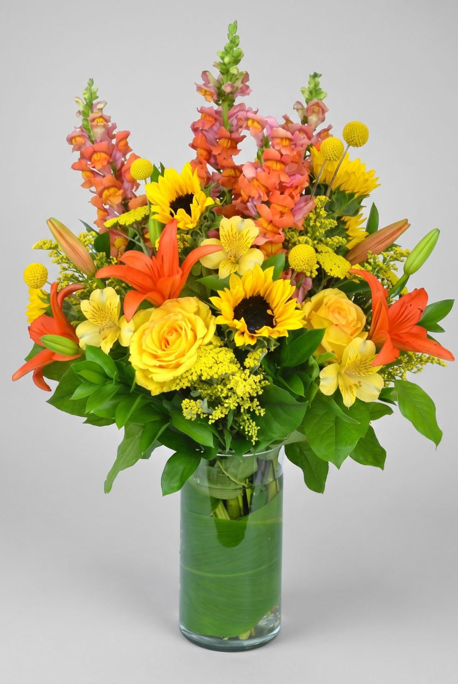 Sunshine Brilliance arrangement of sunflowers, yellow roses, orange lilies, coral snapdragons, yellow alstroemeria and billy balls in a leaf-lined glass cylinder vase