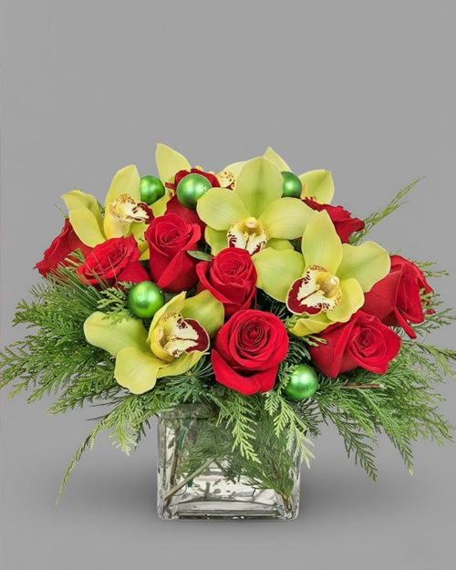 Bouquet of red roses, yellow orchids, and green ornaments in a clear glass vase on a gray background