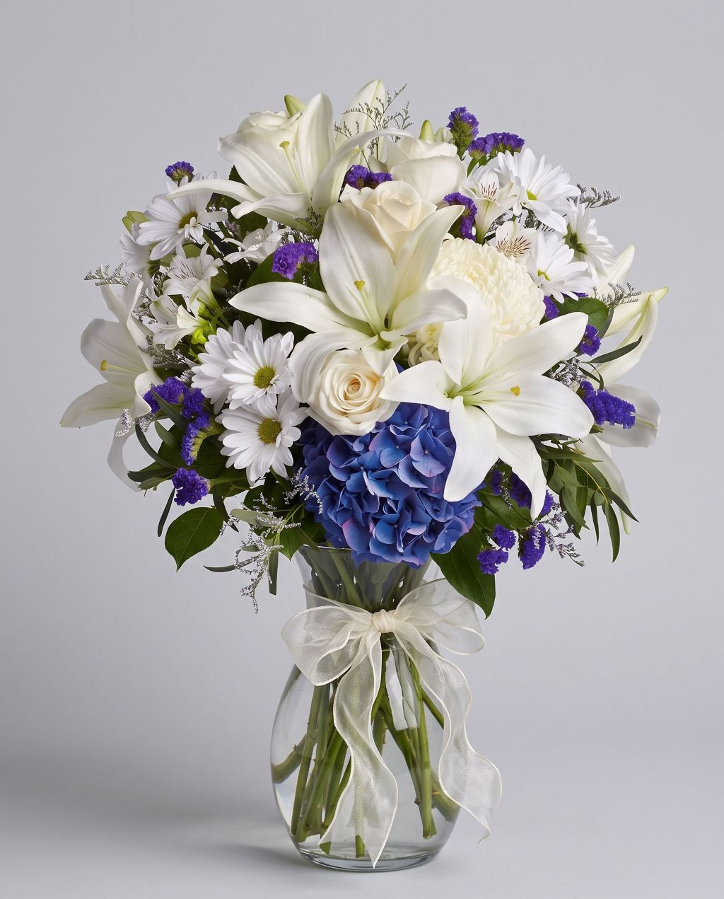 Beautiful in Blue arrangement of blue hydrangea, white lilies, cream roses, white daisies and purple statice in a clear glass vase with an ivory bow