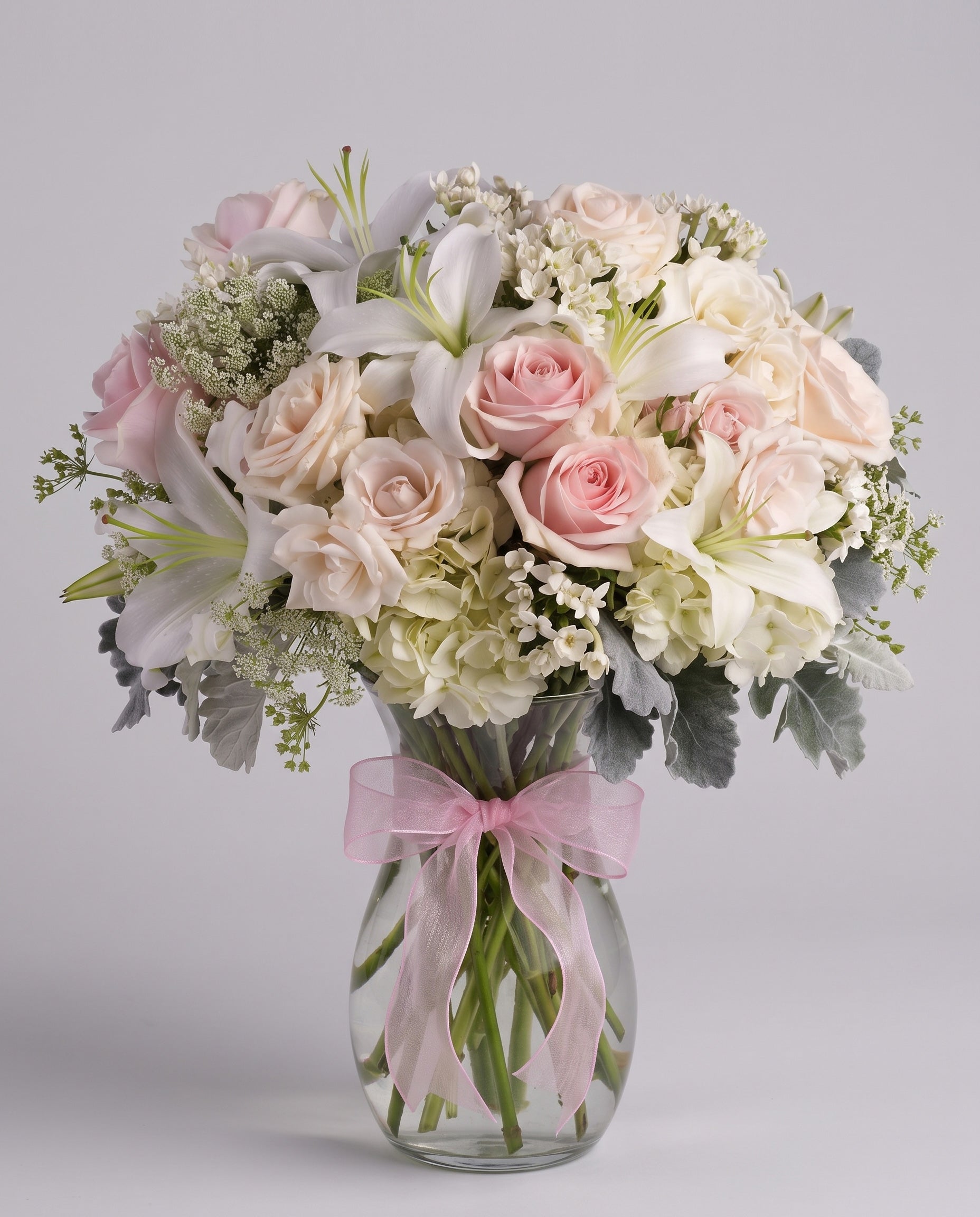 Beautiful Whisper arrangement of blush pink and cream roses, white lilies, white hydrangea and dusty miller in a clear glass vase with a pink bow