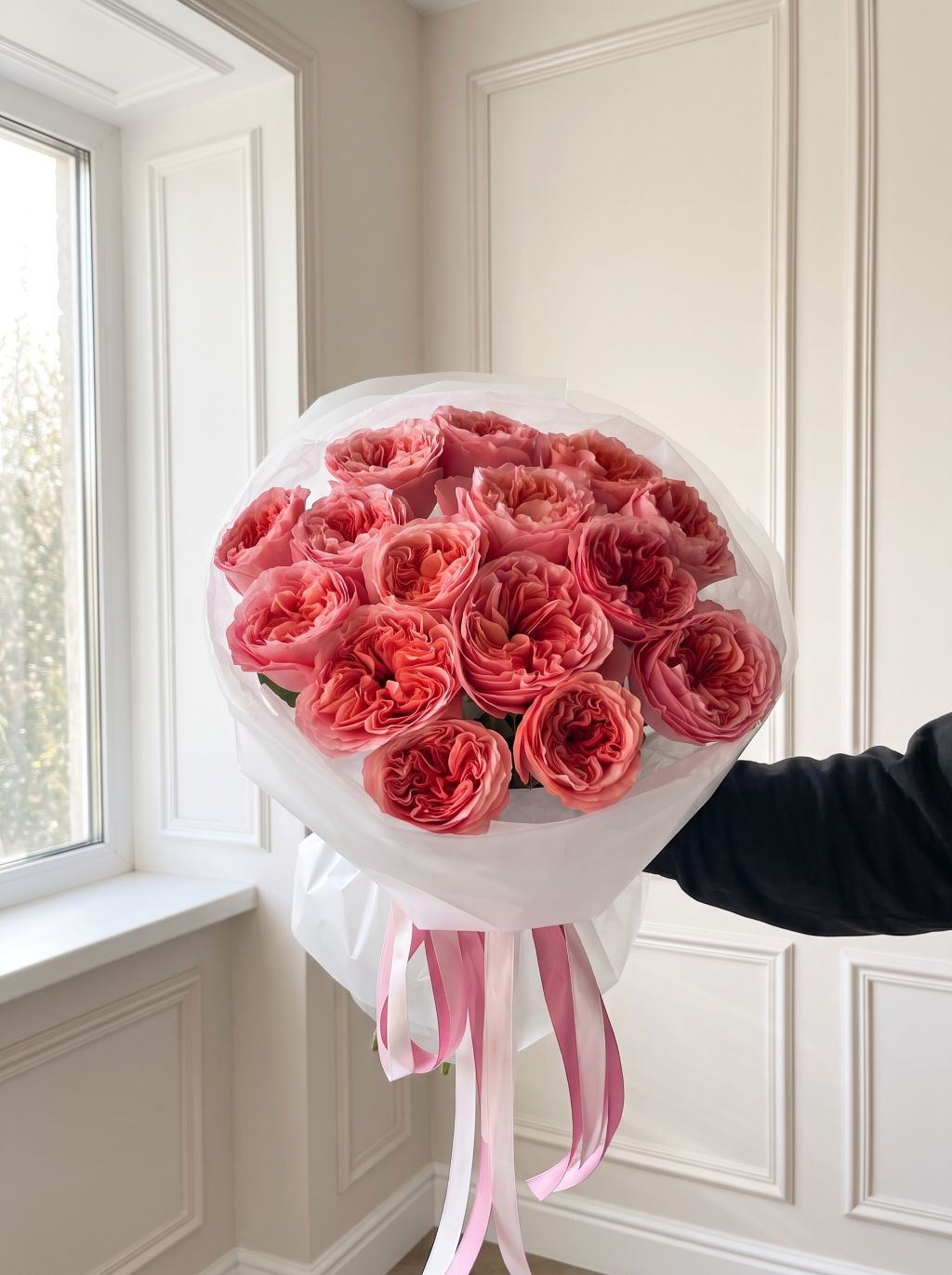 Hand holding a bouquet of coral-pink Candy Xpression garden roses wrapped in white paper with long pink satin ribbons