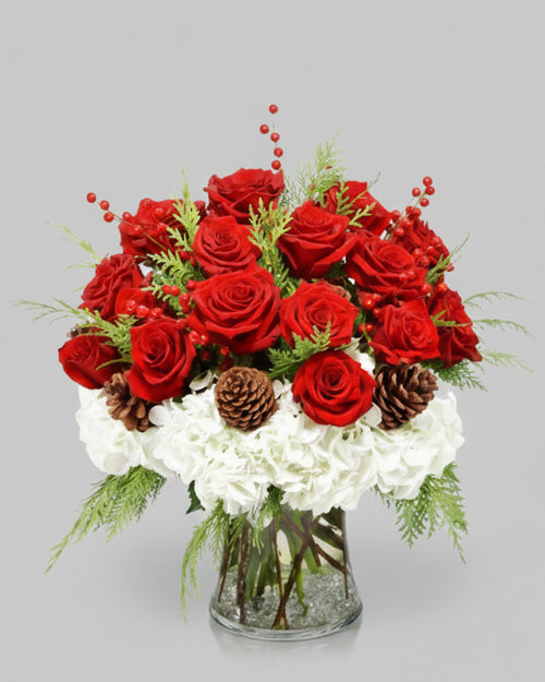 Bouquet of red roses and white flowers with pinecones and berries in a clear vase on a light gray background