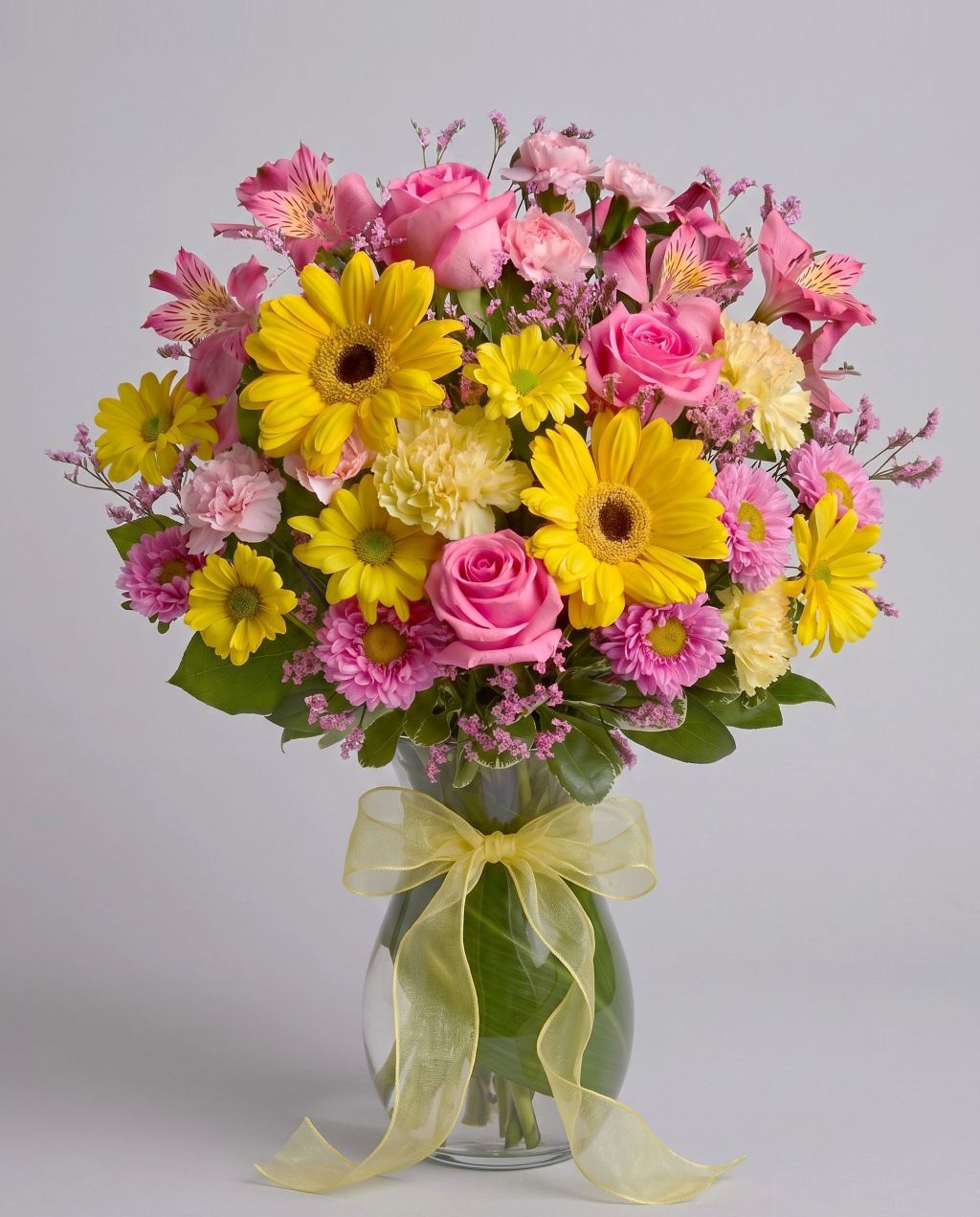 Dazzling Day arrangement of yellow gerbera daisies, pink roses, pink alstroemeria, carnations and asters in a leaf-lined glass vase with a yellow bow