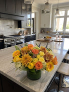 Designer’s choice fall arrangement of orange and yellow roses, bronze cymbidium orchids, yellow gerbera daisies, golden mums and green hydrangea in a leaf-lined glass vase on a kitchen island