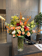 Tall designer’s choice fall arrangement of peach roses, orange calla lilies, green cymbidium orchids, burgundy peonies, orange winterberry and curly willow in a glass cylinder vase on an office reception desk