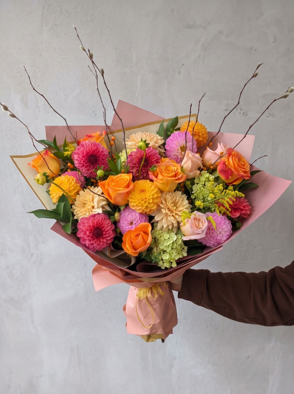 Designer’s choice fall bouquet of orange roses, pink and peach dahlias, green hydrangea, orange berries and pussy willow branches wrapped in blush pink paper