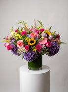 Designer’s choice seasonal arrangement of purple hydrangea, pink and lavender roses, sunflowers, pink anthurium, yellow snapdragons and lilies in a leaf-lined glass vase on a white pedestal