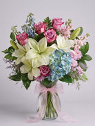 Gentle Comfort arrangement of white lilies, mauve-pink roses, blue hydrangea, blue delphinium, pink stock and purple limonium in a clear glass vase with a pink bow