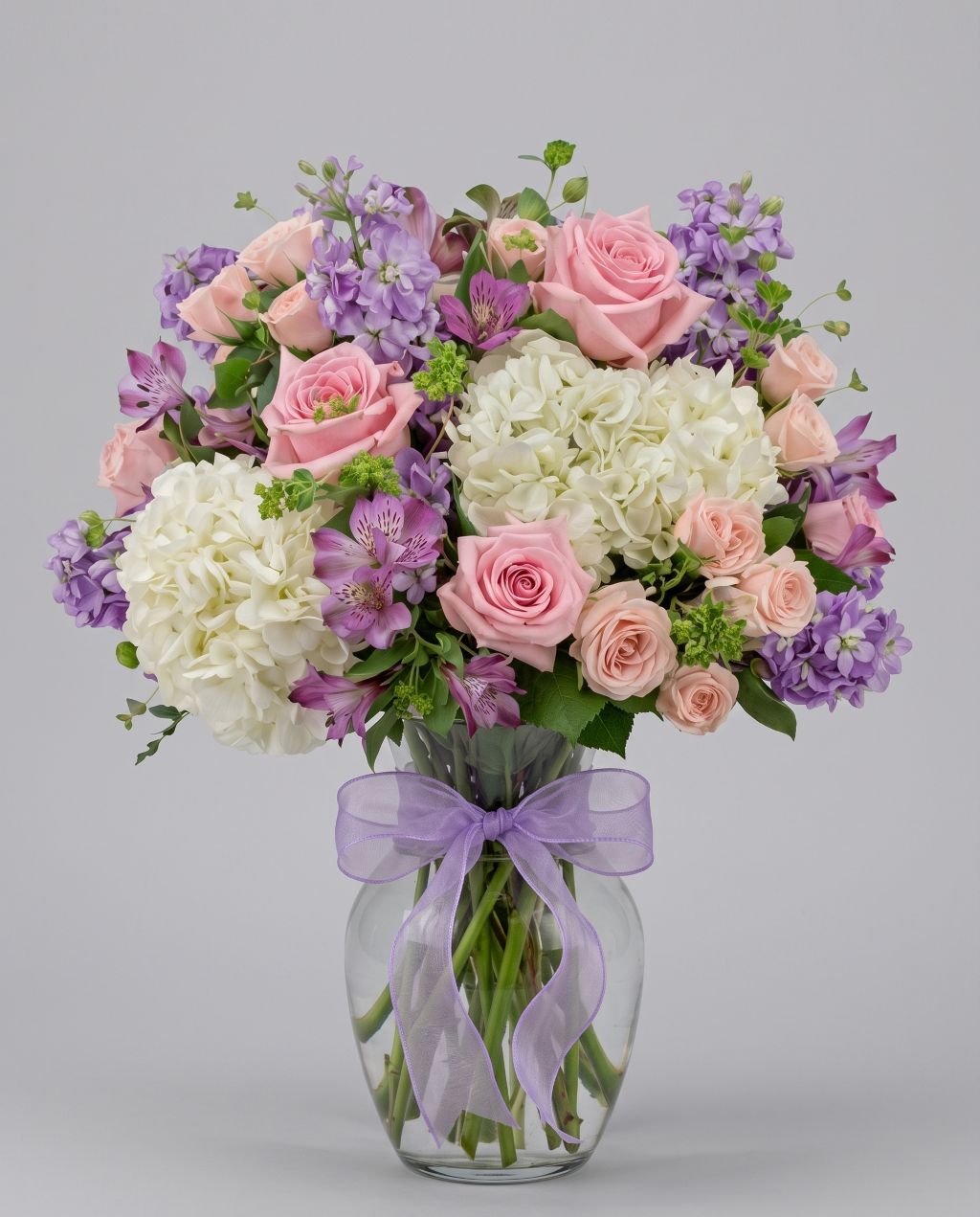Gentle arrangement of pink roses, peach spray roses, white hydrangea, lavender stock and purple alstroemeria in a clear glass vase with a lavender bow