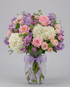 Gentle arrangement of pink roses, peach spray roses, white hydrangea, lavender stock and purple alstroemeria in a clear glass vase with a lavender bow