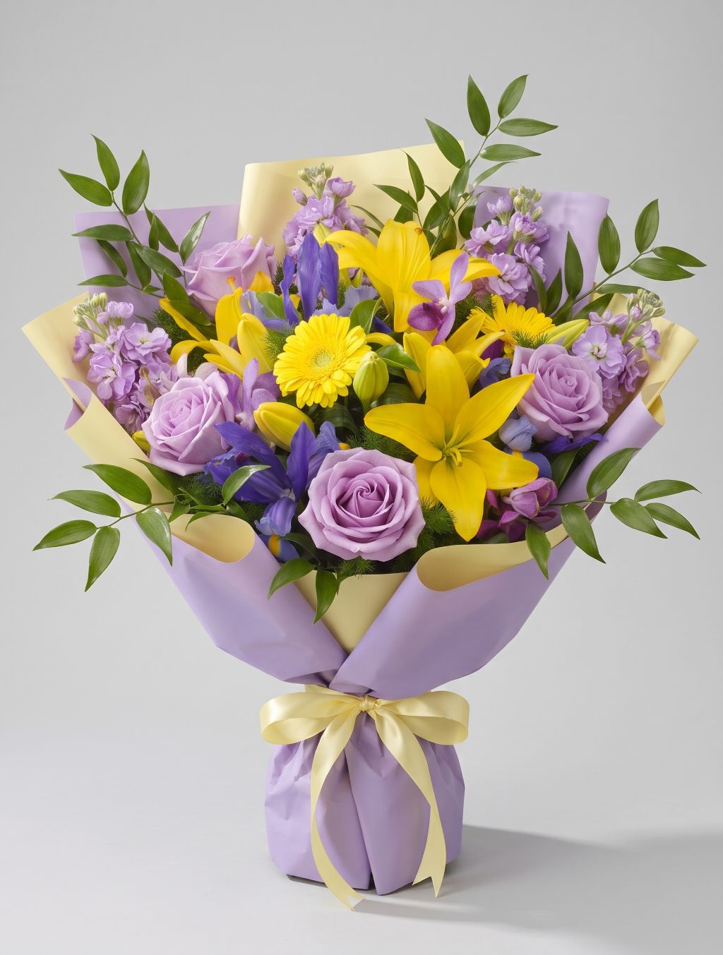 Lavender Sunshine Bouquet of yellow lilies, yellow gerbera daisies, lavender roses, purple iris, lavender stock and purple orchids wrapped in lavender and pale yellow paper with a yellow ribbon