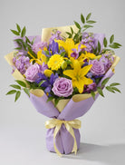 Lavender Sunshine Bouquet of yellow lilies, yellow gerbera daisies, lavender roses, purple iris, lavender stock and purple orchids wrapped in lavender and pale yellow paper with a yellow ribbon