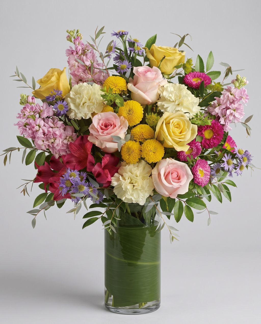 Seasonal spring vase arrangement of pink and yellow roses, cream carnations, yellow button mums, pink stock, red alstroemeria and purple asters in a leaf-lined glass vase