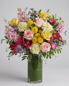 Seasonal spring vase arrangement of pink and yellow roses, cream carnations, yellow button mums, pink stock, red alstroemeria and purple asters in a leaf-lined glass vase
