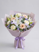 Soft Pastel Bouquet of white lilies, ivory roses, peach gerbera daisies, lavender freesia and statice with eucalyptus wrapped in mauve paper with a lavender ribbon