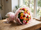 Sunset Fiesta Roses bouquet of mixed red, pink, peach, orange, yellow and white roses wrapped in pink and white paper with a pink ribbon, resting on a wooden table by a sunny window
