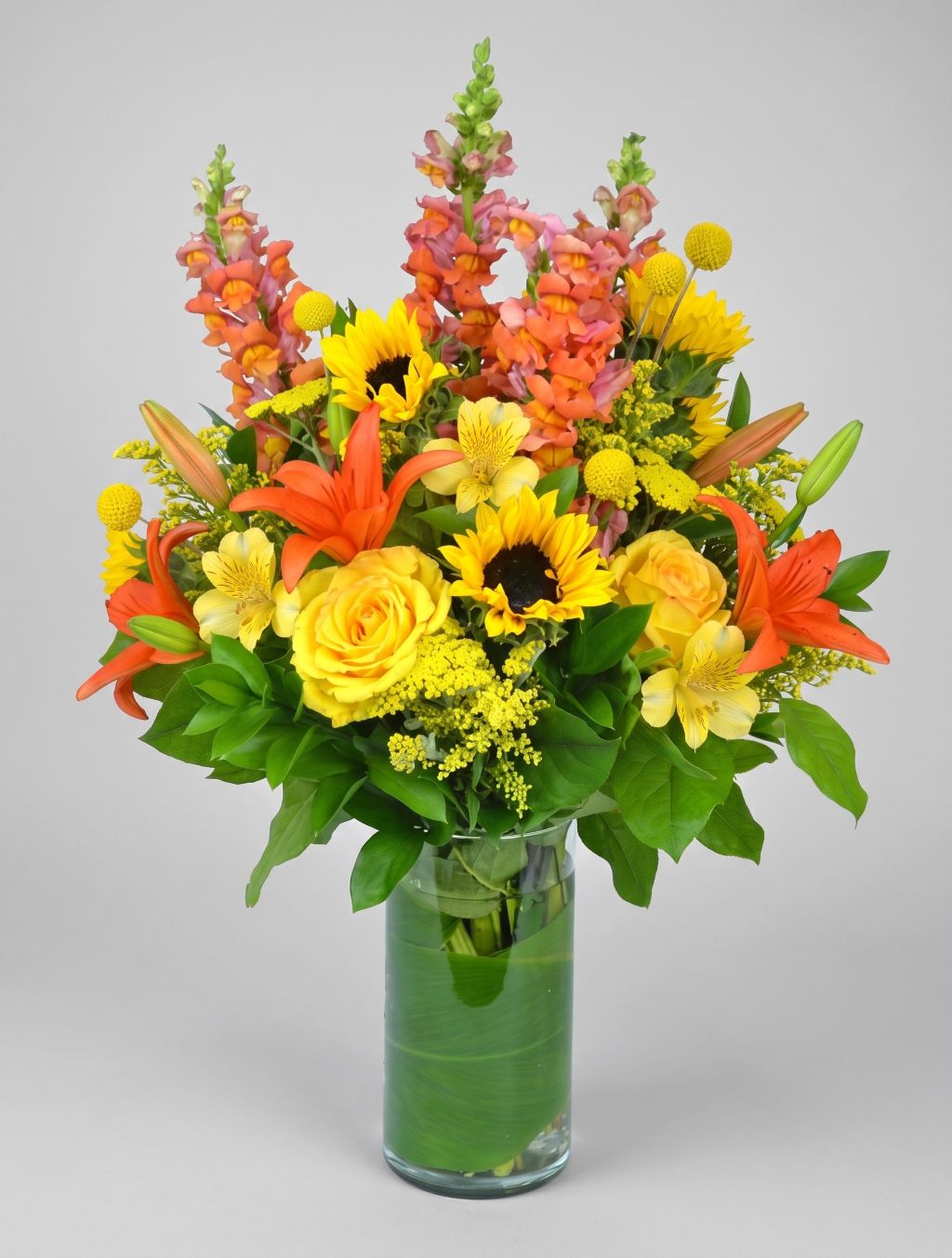 Sunshine Brilliance arrangement of sunflowers, yellow roses, orange lilies, coral snapdragons, yellow alstroemeria and billy balls in a leaf-lined glass cylinder vase