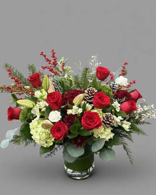 Bouquet of red roses, greenery, and berries in a clear vase on a gray background